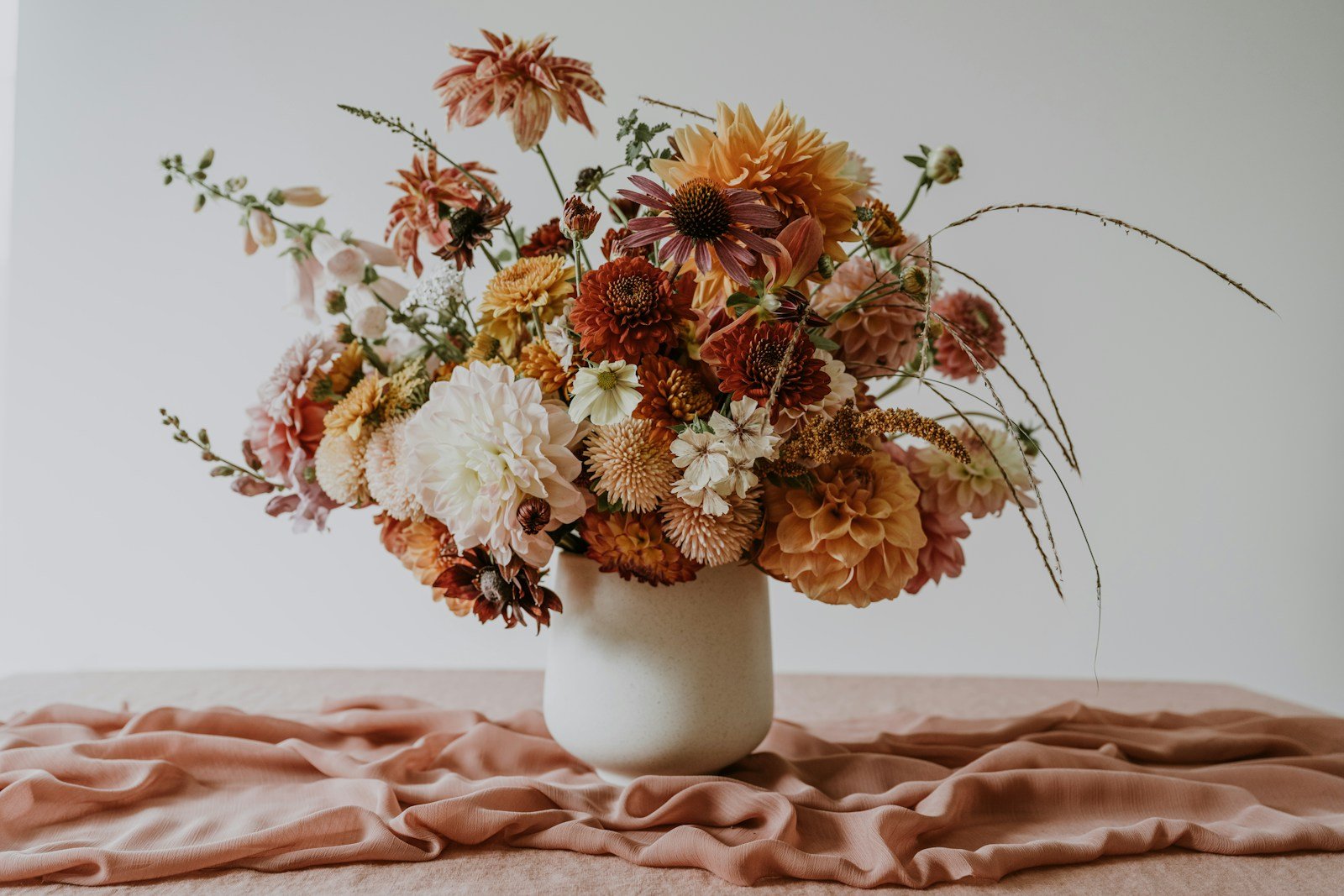 A white vase holds a vibrant bouquet of orange, red, and pink flowers on a draped pink cloth.