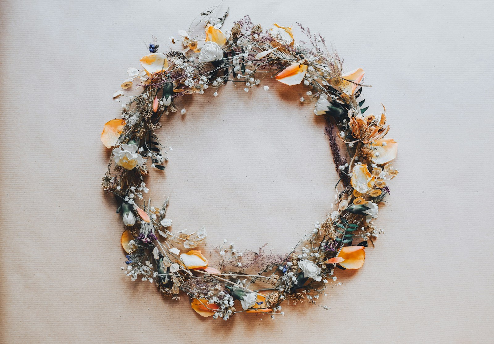 A dried flower wreath with orange petals, white blossoms, and wispy brown foliage on a tan surface.